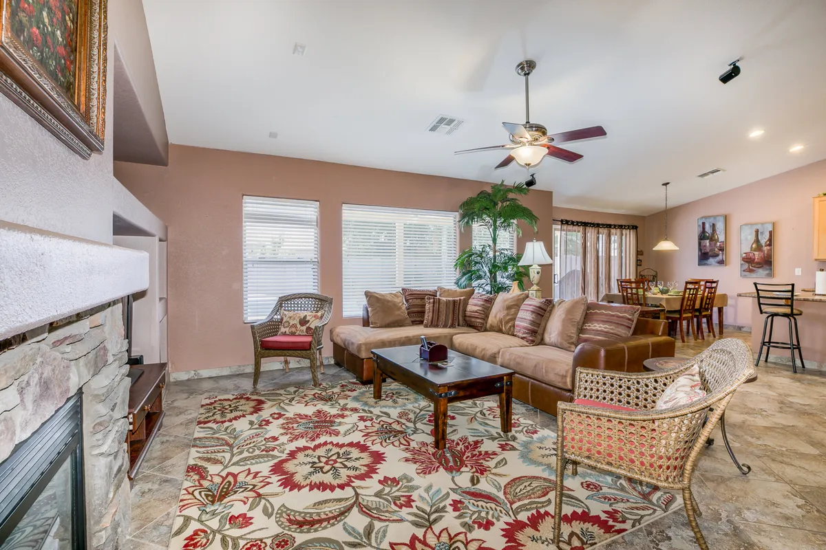 This image captures a spacious great room with a comfortable sectional sofa positioned around an ornate area rug, a dining area with a wooden table, and a fireplace. Large windows in the background allow natural light to pour in, creating a warm, inviting ambiance ideal for family gatherings and relaxation. The open-concept design seamlessly connects the living and dining spaces.