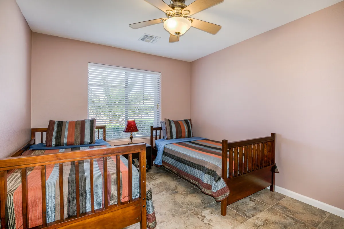 A cheerful bedroom with two wooden twin beds adorned with striped bedding, a window with blinds allowing natural light to filter in, and a tidy, inviting setup. The space is ideal for kids or friends sharing a room.