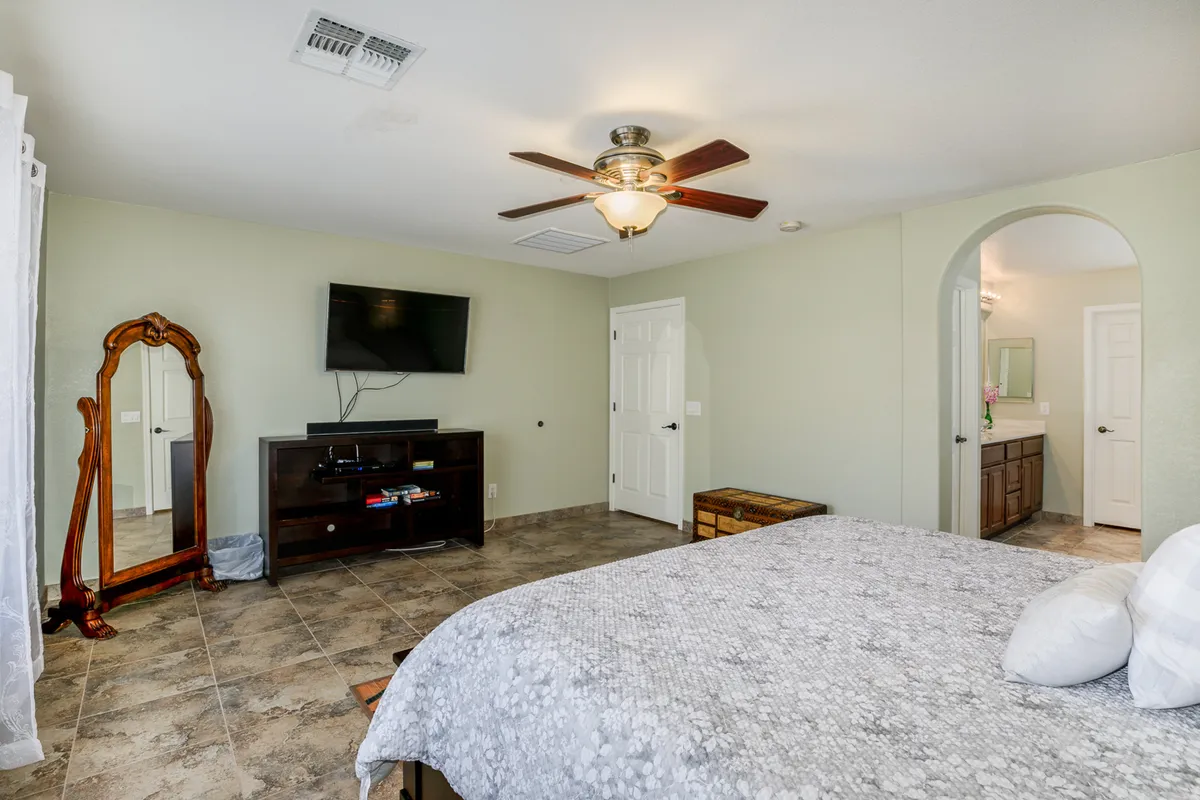 This image shows a comfortable bedroom featuring a plush bed, a wall-mounted TV, a nightstand, a mirror, and a dressing table. The setting is cozy and well-lit, creating a welcoming environment for relaxation or unwinding at the end of the day.