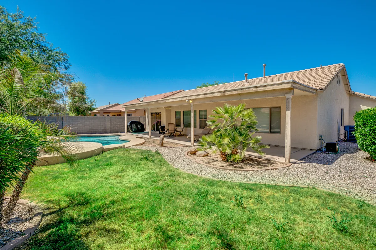 A vibrant backyard scene featuring a sparkling swimming pool, comfortable patio seating, a landscaped garden, and part of the house exterior, all under clear, sunny skies. The area looks spacious and welcoming, perfect for both relaxation and family activities.