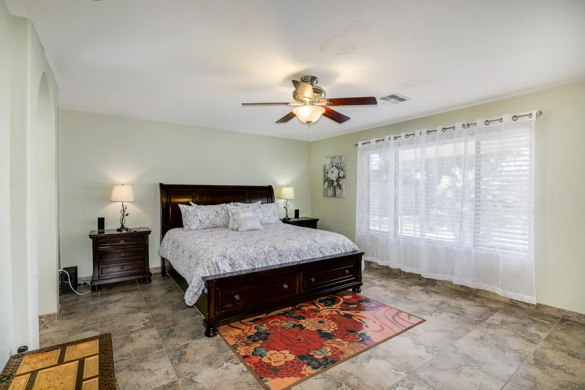 A serene bedroom scene with a neatly made bed, a large window letting in soft daylight, a cozy rug, and matching nightstands. The atmosphere feels peaceful and restorative, ideal for restful sleep or a relaxing nap.