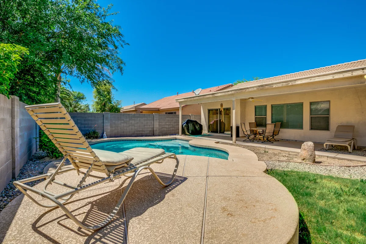 A backyard scene showing a sparkling swimming pool, a cozy lounge chair, and comfortable patio seating. This inviting setup is perfect for sunbathing, casual conversation, or enjoying a meal outdoors. The tranquil mood and thoughtful design invite relaxation and socializing.