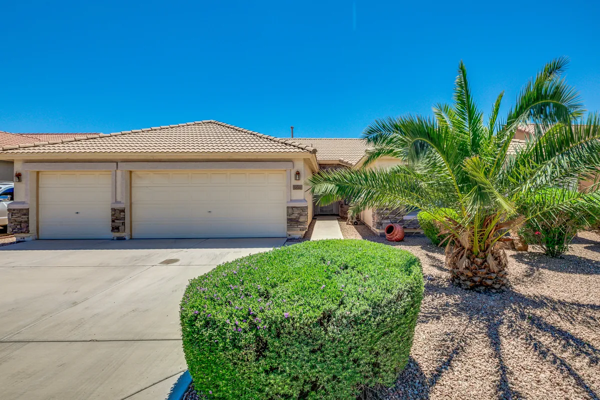 A wide exterior view of the home's front, prominently featuring the two-car garage, a lush palm tree, manicured shrubs, and a rock landscape. The garage doors are clearly visible, making it easy to visualize convenient parking options.