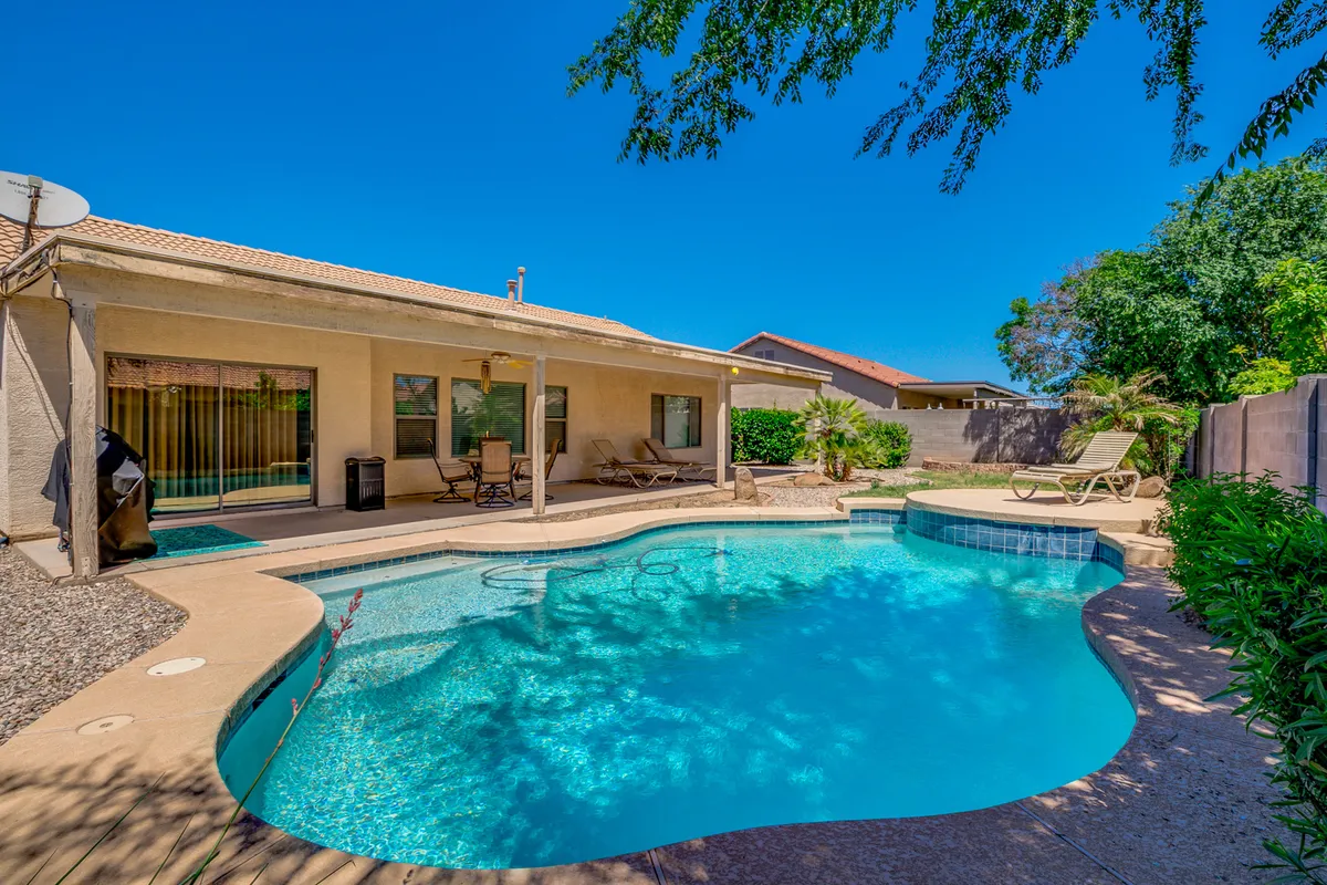 A sun-filled backyard featuring a sparkling swimming pool, a spacious patio area, and lush green landscaping. Lounge chairs are positioned poolside, and the patio provides ample space for both relaxing and outdoor dining. The vibrant greenery and open sky create an inviting oasis atmosphere.