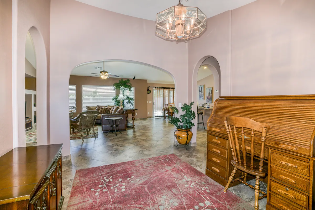 A dedicated workspace with a wooden desk, an elegant chandelier overhead, an area rug, and arched doorways in the background. The space looks quiet and conducive to focused work or planning.