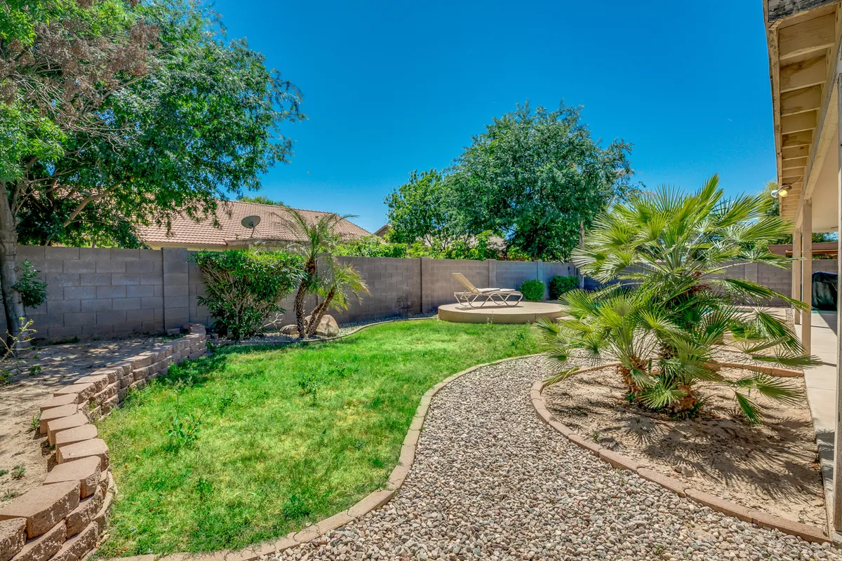 A lush backyard lawn with a decorative gravel pathway runs alongside a lounge chair and palm trees. The space is surrounded by a privacy wall and features a serene spot for play, relaxation, and gatherings. The greenery and open area are ideal for pets and kids alike.